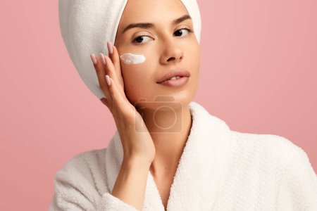 Photo for Crop young female model in white bathrobe and towel wrapped around head applying moisturizing cream on face and looking away against pink background - Royalty Free Image
