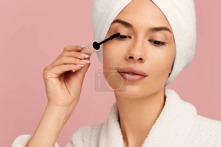 Téléchargez les photos : Belle jeune femme appliquant le mascara sur les cils avec une serviette autour de la tête après la douche sur fond rose - en image libre de droit