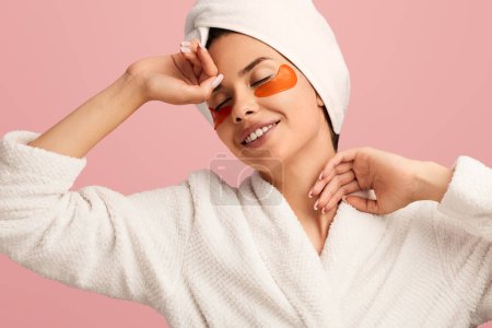 Téléchargez les photos : Joyeux jeune femme en peignoir et serviette sur la tête avec des patchs touchant le front et le cou, tout en s'étirant en studio sur fond rose pendant la routine de soins de la peau - en image libre de droit