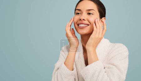 Téléchargez les photos : Femme sensuelle souriante en peignoir blanc touchant les joues tout en appliquant un produit hydratant sur le visage avec une peau parfaite sur fond turquoise - en image libre de droit