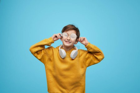 Photo for Astonished boy adjusting stylish glasses and looking at camera against blue background - Royalty Free Image