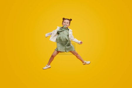 Photo for Full body cute kid in stylish dress swinging arms and smiling while jumping against bright yellow background - Royalty Free Image