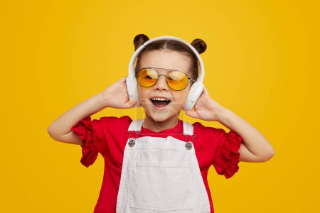 Téléchargez les photos : Douce petite fille en tenue élégante lumineuse écoutant de la musique dans les écouteurs et regardant étonné de la caméra sur fond jaune - en image libre de droit