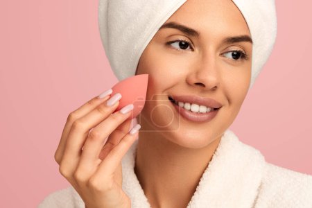 Téléchargez les photos : Gros plan de Happy jeune femme souriant et utilisant l'éponge de maquillage comme routine cosmétique après la douche sur fond rose - en image libre de droit