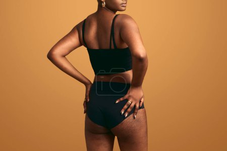 Photo for Back view of crop African female with imperfect body showing buttocks while wearing black lingerie on brown background - Royalty Free Image