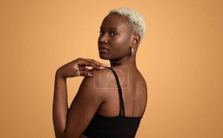 Téléchargez les photos : Jeune modèle féminin afro-américain pensif en haut noir avec les cheveux courts regardant par-dessus l'épaule à la caméra sur fond beige en studio - en image libre de droit