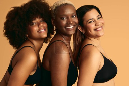 Téléchargez les photos : Jeunes filles heureuses et diversifiées en lingerie noire avec différentes couleurs de peau debout près et souriant sur fond orange regardant la caméra - en image libre de droit