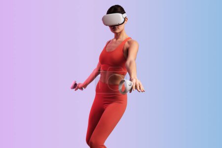 Photo for Young fit lady in red sportive outfit and VR headset holding controllers, while playing videogame interacting with invisible reality spending leisure time against gradient background - Royalty Free Image