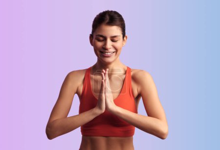 Photo for Positive young female athlete in red top doing yoga while meditating with hands in namaste gesture and closed eyes against gradient background - Royalty Free Image