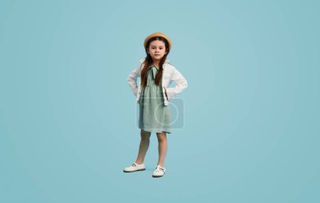 Téléchargez les photos : Adorable petit enfant en robe élégante et chapeau regardant loin tout en se tenant debout sur fond bleu - en image libre de droit