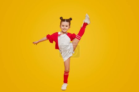 Téléchargez les photos : Adorable petite fille en tenue lumineuse soulevant la jambe et souriant tout en dansant sur fond jaune. Cours de danse pour enfants concept - en image libre de droit