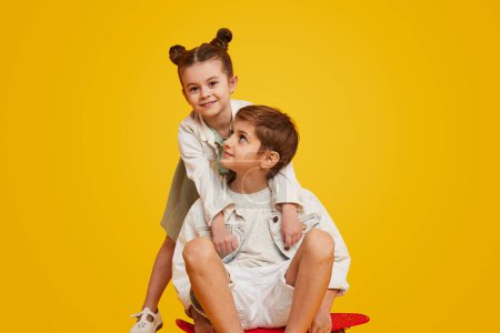 Photo pour Cool garçon à la mode avec longboard embrassant adorable petite fille dans une tenue élégante et regardant ensemble la caméra sur fond jaune - image libre de droit
