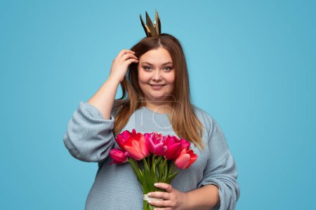 Téléchargez les photos : Belle plus la taille femelle en papier couronne tenant tas de tulipes mignonnes et souriant tout en se tenant debout sur fond bleu et en regardant la caméra - en image libre de droit