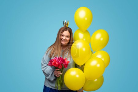 Foto de Emocionado hembra regordeta en la corona de papel que sostiene ramo de tulipanes rojos y racimo de globos amarillos mientras celebra el cumpleaños sobre fondo azul - Imagen libre de derechos