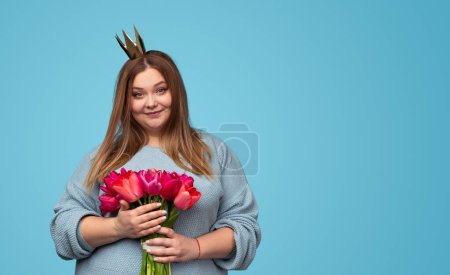 Photo for Charming plus size woman in paper crown holding bouquet of tulips and looking at camera while standing on blue background - Royalty Free Image