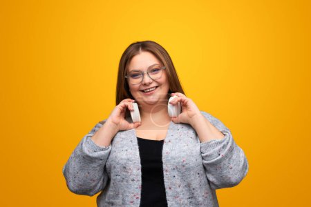 Foto de Emocionada hembra de tamaño grande con auriculares modernos sonriendo mientras está de pie sobre fondo amarillo - Imagen libre de derechos
