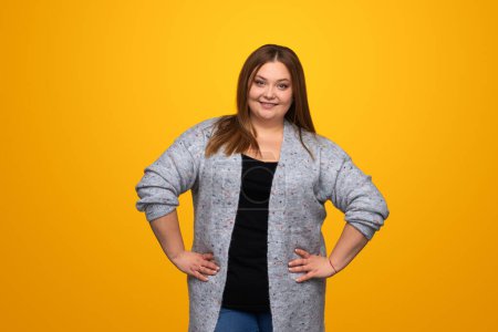 Foto de Mujer joven con sobrepeso positivo en traje de moda manteniendo las manos en la cintura y mirando a la cámara mientras está de pie sobre fondo amarillo - Imagen libre de derechos