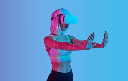 Foto de Mujer futurista joven en peluca y casco VR tocando la pantalla invisible en realidad virtual mientras está de pie bajo la luz de neón sobre fondo azul - Imagen libre de derechos