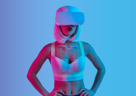 Foto de Mujer joven futurista en sujetador y gafas VR explorando la realidad virtual mientras está de pie bajo la luz de neón sobre el fondo azul - Imagen libre de derechos
