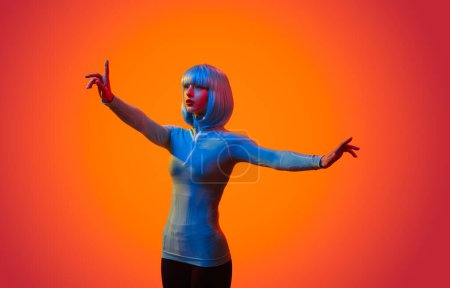 Foto de Joven hembra futurista en ala levantando el brazo y tocando la pantalla invisible con el dedo de pie en el estudio sobre fondo naranja - Imagen libre de derechos