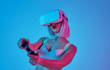 Téléchargez les photos : Jeune femme auto-assurée aux cheveux blonds et aux lèvres rouges jouant au jeu vidéo avec joystick dans un casque VR moderne en studio néon bleu - en image libre de droit