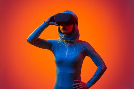 Photo for Young female in VR goggles wearing futuristic clothes with blue hair against neon orange background - Royalty Free Image