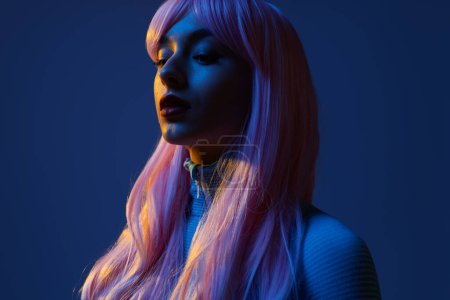 Téléchargez les photos : Vue latérale du jeune modèle féminin futuriste confiant avec de longs cheveux roses sur fond bleu - en image libre de droit