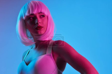 Téléchargez les photos : Jeune femme futuriste portant soutien-gorge et perruque debout dans un studio éclairé par des néons sur fond bleu détournant les yeux - en image libre de droit