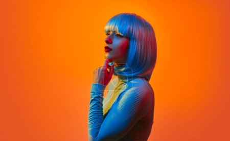 Téléchargez les photos : Vue latérale de la jeune femme en tenue futuriste à la mode touchant le menton et regardant loin tout en se tenant debout sur fond orange brillant - en image libre de droit