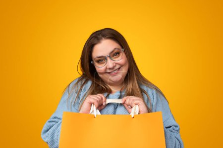 Foto de Sonriendo más tamaño joven hembra en gafas mirando a la cámara mientras está de pie con bolsas de papel en las manos contra el fondo amarillo - Imagen libre de derechos