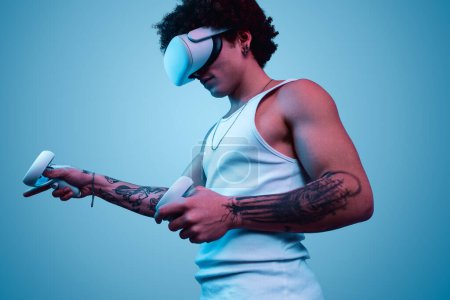 Téléchargez les photos : Vue latérale du mâle afro-américain moderne en lunettes VR et avec des contrôleurs jouant à un jeu vidéo sur fond bleu - en image libre de droit