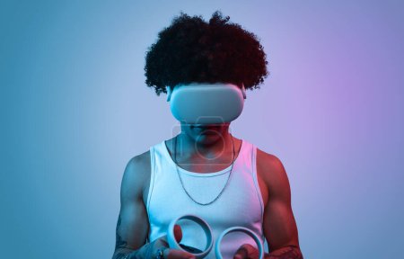 Photo for Young ethnic male in VR goggles and with controllers playing video game against neon light with blue and purple background - Royalty Free Image
