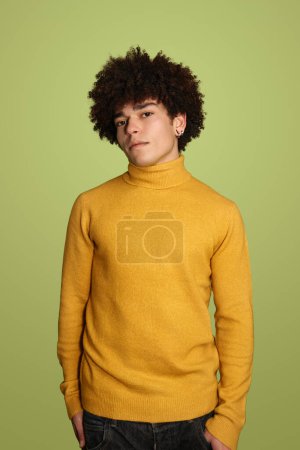 Foto de Joven macho hispano con el pelo rizado usando suéter amarillo y mirando a la cámara mientras está de pie sobre fondo verde - Imagen libre de derechos