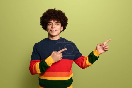 Photo for Happy ethnic male with curly hair pointing aside while standing against green background and looking at camera - Royalty Free Image