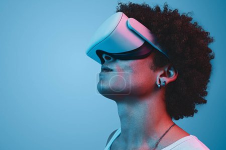 Téléchargez les photos : Vue latérale d'un homme afro-américain sérieux portant des lunettes VR et explorant le cyberespace sur fond bleu en studio sous la lumière du néon - en image libre de droit