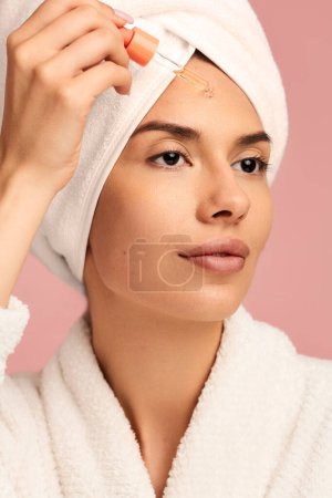 Téléchargez les photos : Jeune femme confiante avec une peau parfaite en peignoir blanc et serviette sur la tête appliquer de l'huile sérique sur le visage et détourner les yeux, lors de la procédure de beauté quotidienne sur fond rose - en image libre de droit