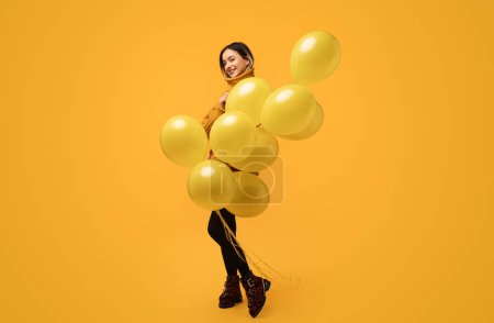 Photo for Full body side view of young female holding bunch of balloons while standing against yellow background - Royalty Free Image