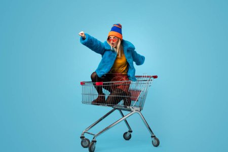 Photo for Side view of crazy trendy young female in bright clothes holding paper bags and riding shopping cart in superhero pose against blue background - Royalty Free Image