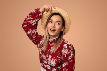 Photo for Surprised young woman in fashionable floral dress adjusting straw hat and looking away while standing against beige background - Royalty Free Image
