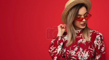 Foto de Elegante joven dama en tonos llamativos y sombrero mirando hacia otro lado mientras está de pie sobre un fondo rojo vivo con espacio para copiar - Imagen libre de derechos