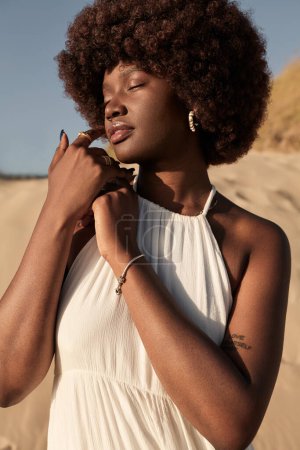 Téléchargez les photos : Jeune mannequin africaine douce avec des cheveux afro dans des vêtements à la mode touchant le visage avec le doigt, tout en se tenant à la campagne par une journée ensoleillée avec les yeux fermés - en image libre de droit