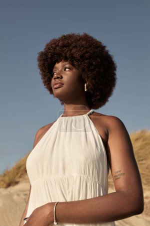 Foto de Bajo ángulo de mujer africana joven en vestido blanco con tatuaje en el brazo, de pie en la naturaleza en el día de verano soleado contra el cielo azul sin nubes y mirando hacia otro lado - Imagen libre de derechos