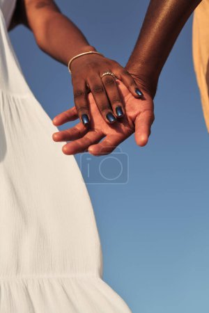 Téléchargez les photos : Angle bas de la culture anonyme femme noire portant une robe blanche tenant la main du mari contre le ciel bleu sans nuages le jour d'été en plein soleil - en image libre de droit