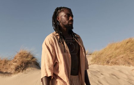 Téléchargez les photos : Jeune homme africain en vêtements beiges avec barbe debout près de l'herbe sèche contre ciel bleu sans nuages le jour ensoleillé et détournant le regard pensivement - en image libre de droit