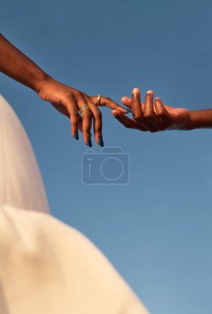 Téléchargez les photos : Cultiver l'homme africain anonyme toucher doucement doigt de petite amie portant des anneaux contre ciel bleu sans nuages près doux tissu ondulant beige dans la lumière du soleil - en image libre de droit