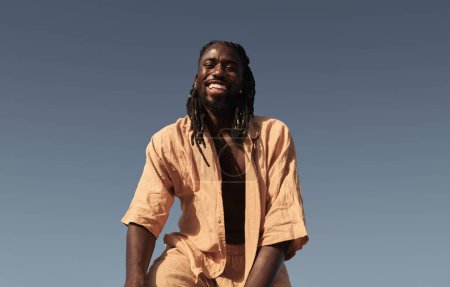 Foto de Bajo ángulo de hombre africano feliz en ropa de verano casual mirando a la cámara con sonrisa dentada mientras está de pie contra el cielo azul sin nubes a la luz del sol - Imagen libre de derechos