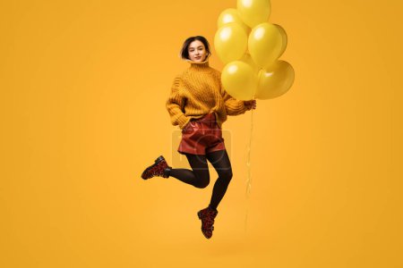 Foto de Cuerpo completo freakish joven dama en colorido traje celebración de racimo de vivos globos y saludo mientras salta sobre fondo amarillo - Imagen libre de derechos