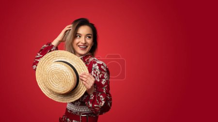 Téléchargez les photos : Jeune femme charnue dans des vêtements floraux élégants tenant un chapeau de paille et regardant l'espace de copie sur fond rouge vif - en image libre de droit