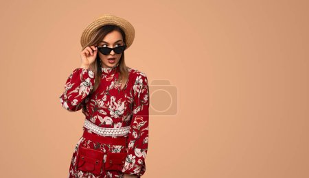 Foto de Mujer sorprendida en ropa de moda con patrón floral con gafas de sol y sombrero de paja mirando hacia otro lado con interés - Imagen libre de derechos
