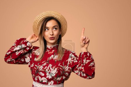 Foto de Sorprendido joven dama en vestido floral de moda apuntando y mirando hacia arriba mientras se ajusta sombrero de paja sobre fondo beige - Imagen libre de derechos